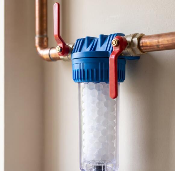 Een polyfosfaatdoseerfilter met witte korrels, blauwe kop en rode afsluitkranen op een koperen leiding